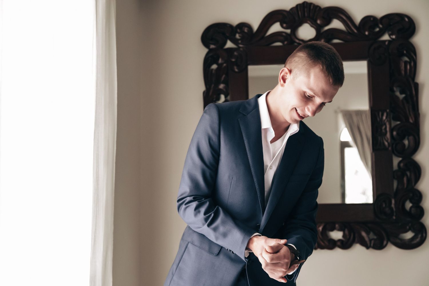 Handsome groom in navy suit adjusting cufflinks near ornate mirror before wedding.