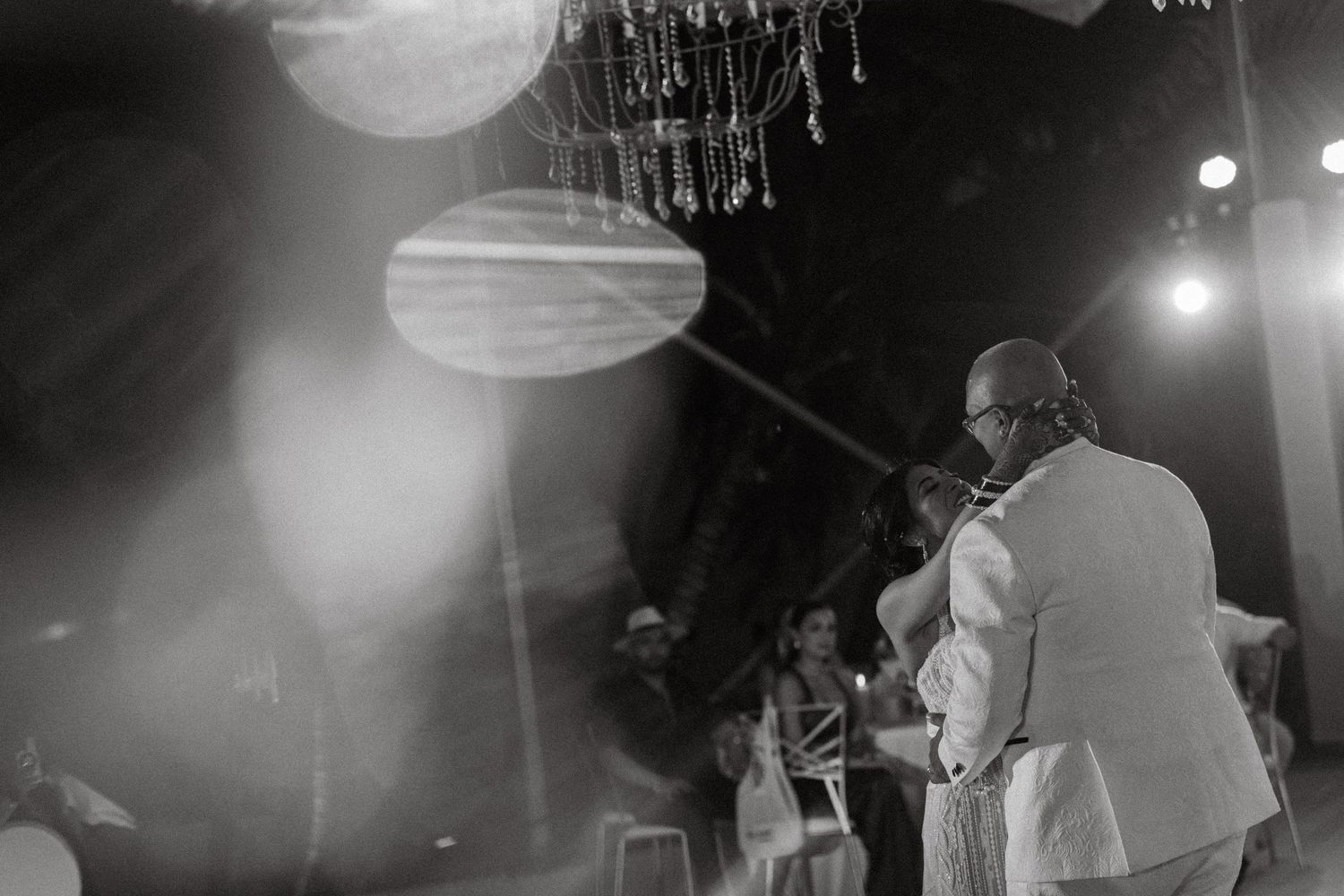 Black and white photo of groom in white suit during outdoor wedding ceremony under decorative chandelier.