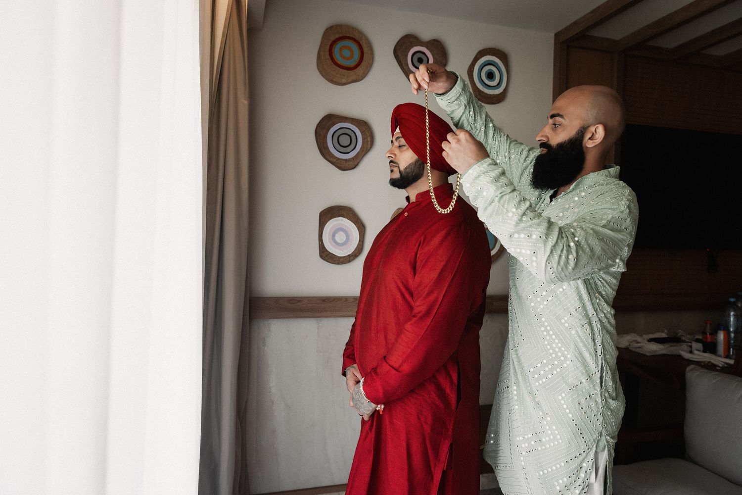 A man in green kurta helps another in red sherwani tie a turban during a traditional South Asian wedding preparation.
