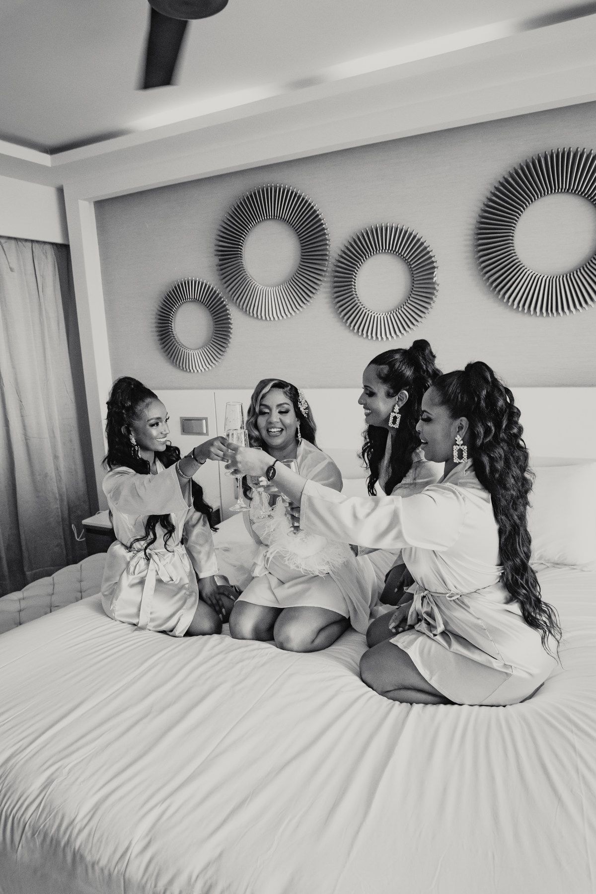 Four bridesmaids in white robes toasting with champagne on a bed in a hotel room.