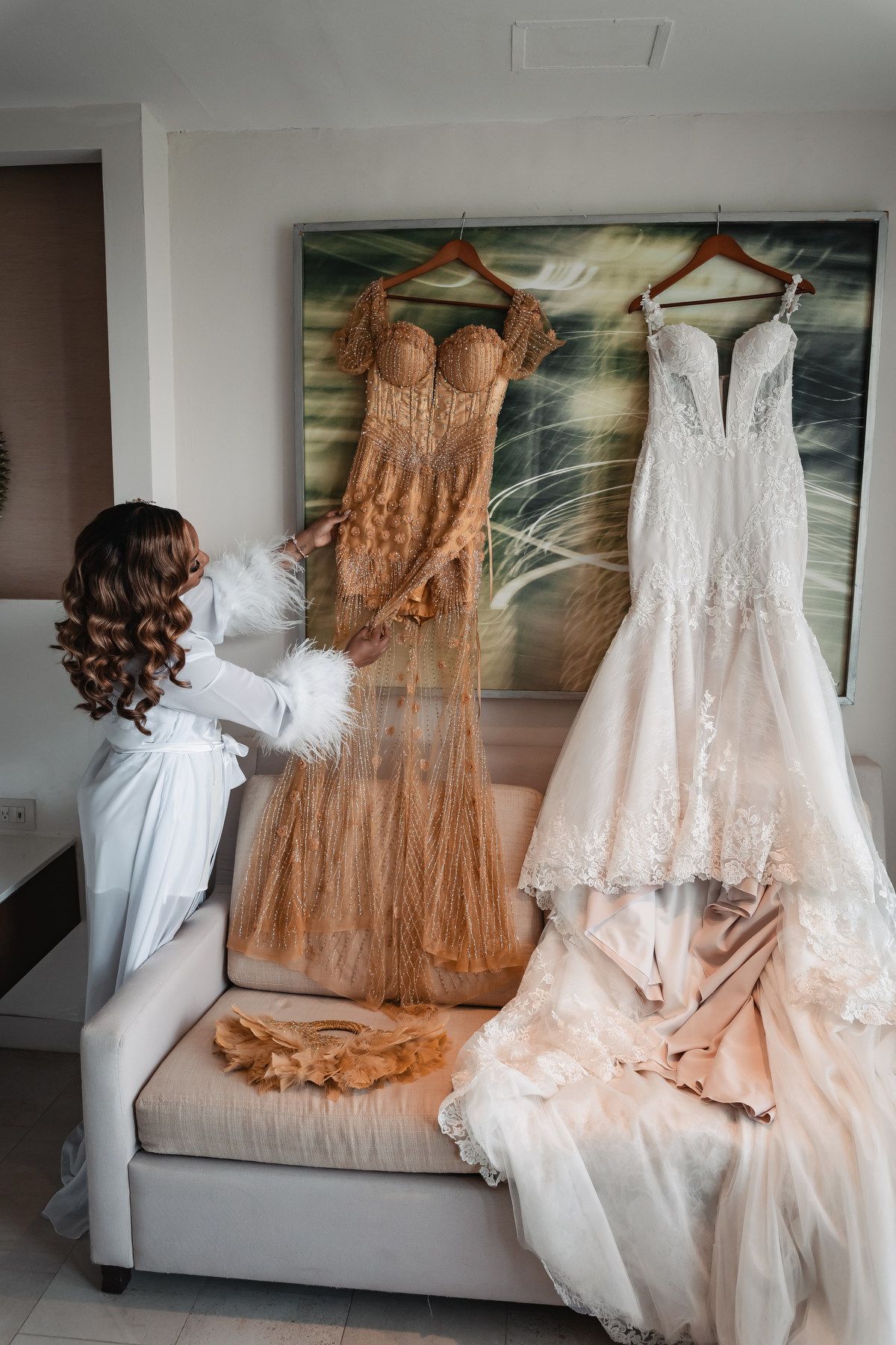 Bride in white robe examines elegant gold and white wedding gowns hanging in a hotel room.