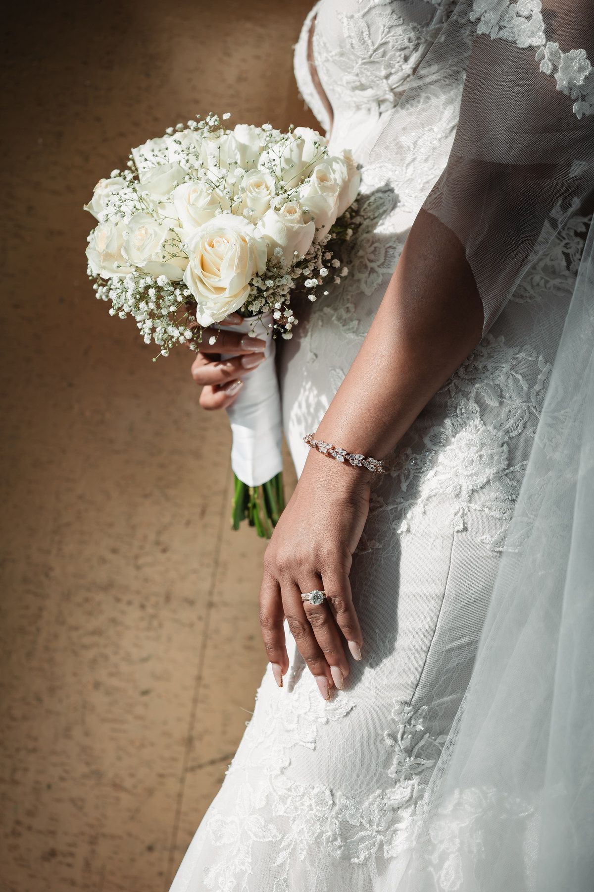 Bride in lace gown holding white rose bouquet displaying diamond ring and bracelet.