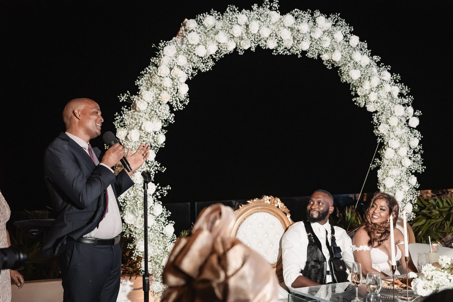 Man giving wedding toast under floral arch while bride and groom listen at reception.