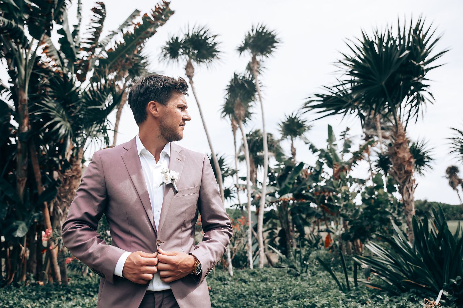 Groom in pink suit poses among tropical palm trees at outdoor venue.