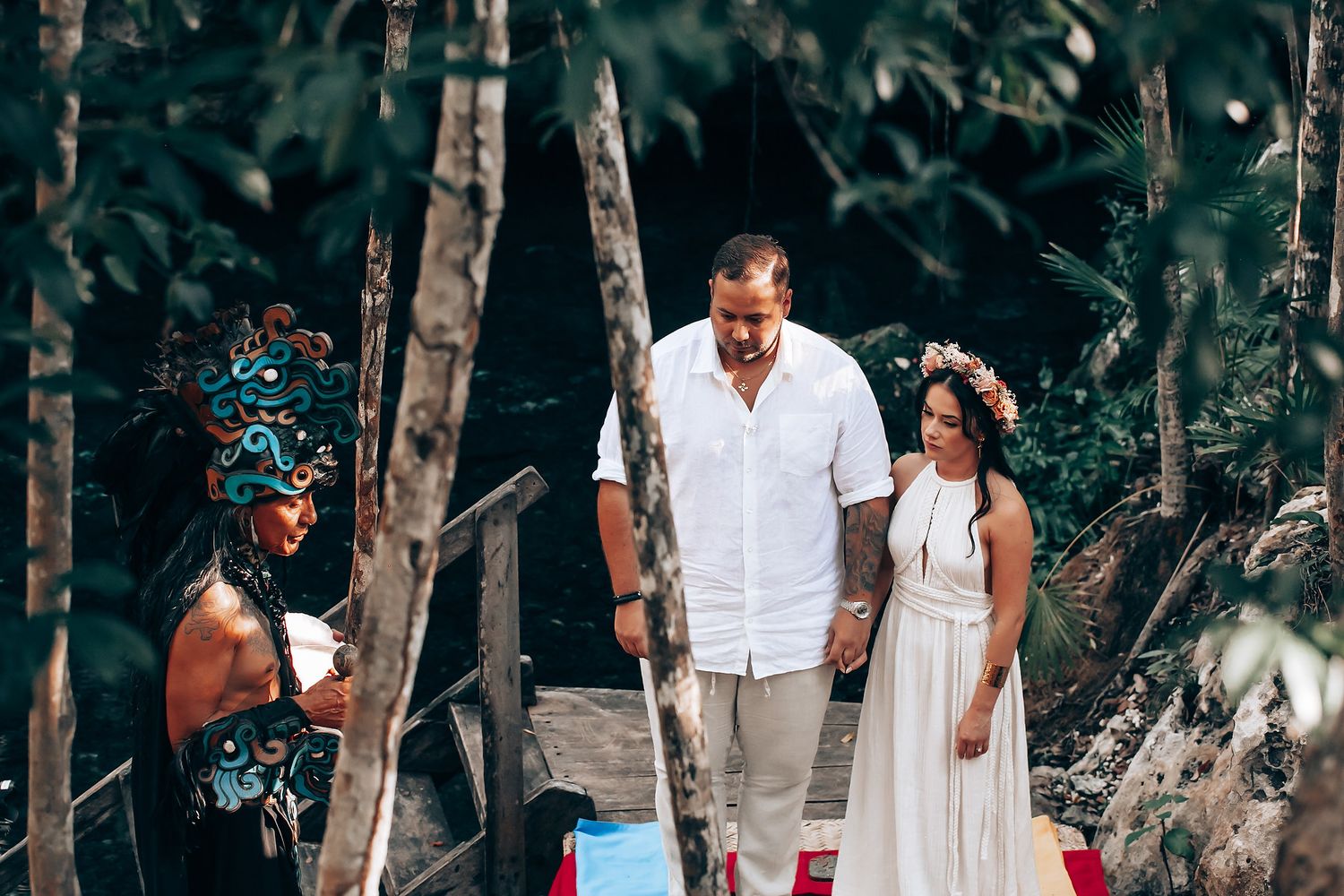 A romantic outdoor photoshoot features a couple in white clothing walking among tall trees in a forest setting.