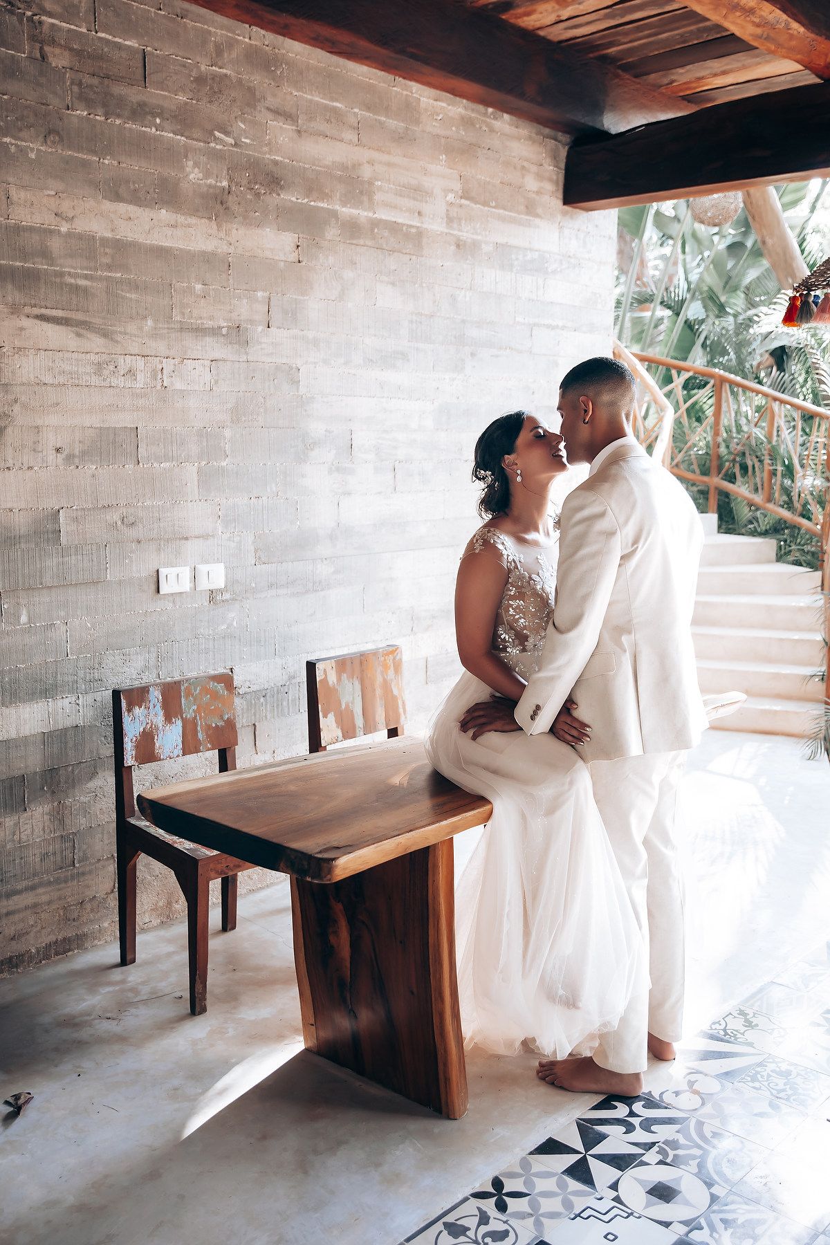 A romantic wedding moment against a concrete wall with wooden chairs and natural lighting.
