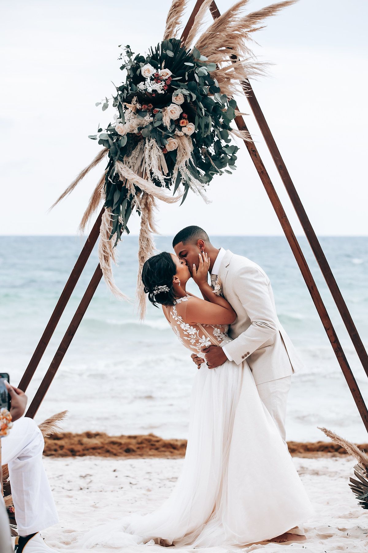 Romantic beach wedding ceremony under wooden arch decorated with pampas grass and flowers.