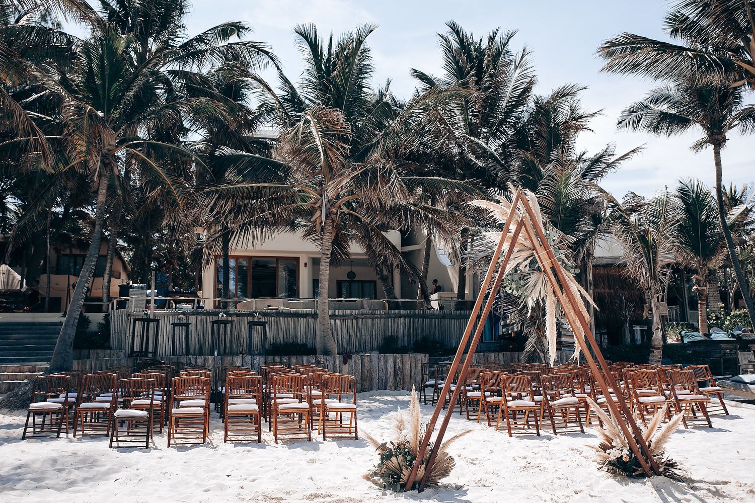 Wooden chairs arranged on a sandy beach with palm trees and a bamboo arch for a tropical beach wedding ceremony.