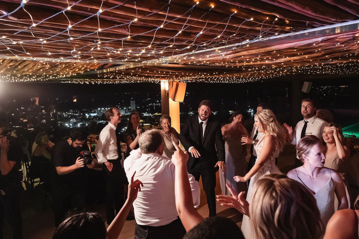 Wedding guests dancing and celebrating under string lights at an indoor reception.