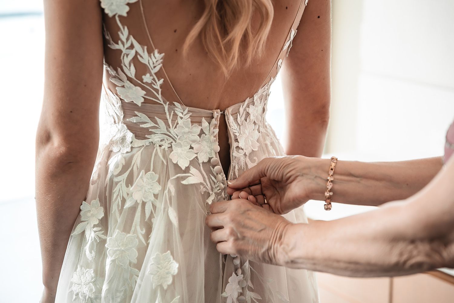 Close-up details of a delicate white wedding dress with floral lace appliques being fastened.