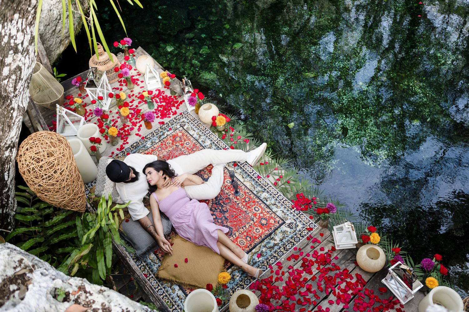 Couple lying on a floral decorated rug by a serene pond, surrounded by rose petals and lush greenery.