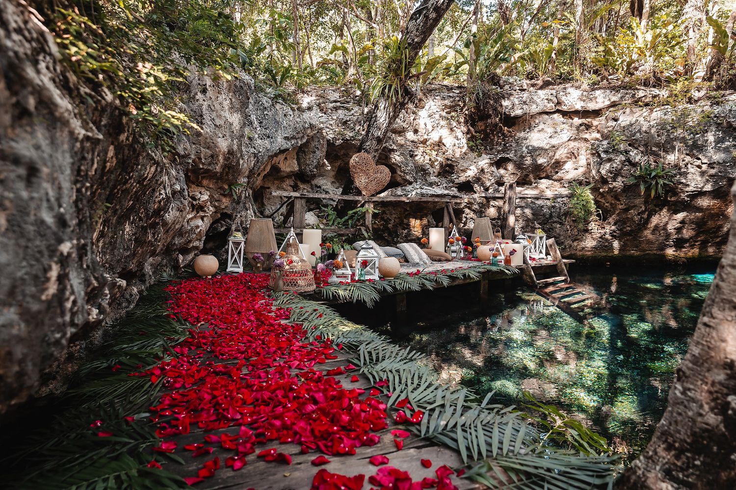 Romantic cenote setup with red rose petals floating on crystal-clear water surrounded by lush jungle rock formations.