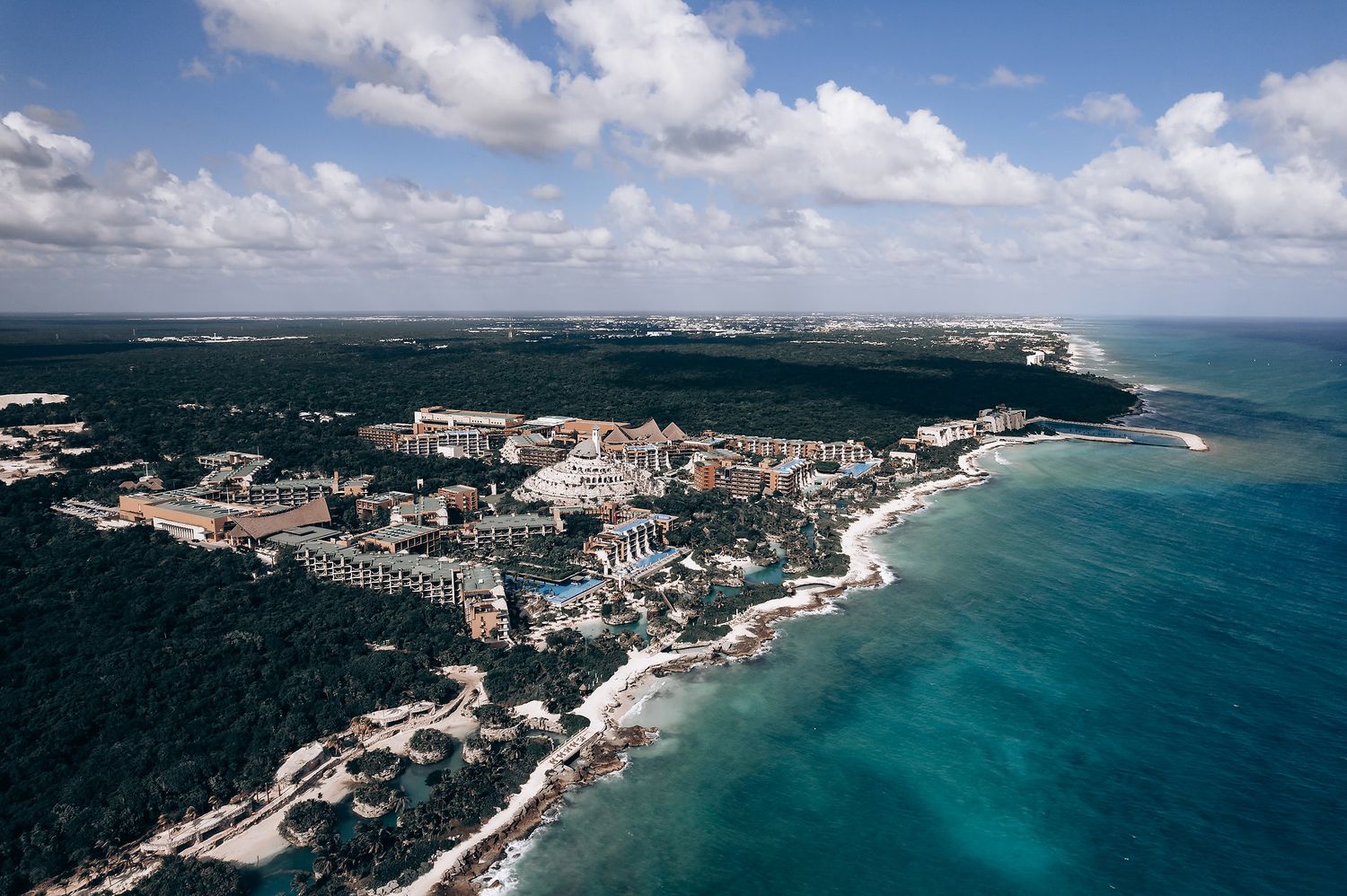 Aerial panoramic view of white cliffs along a coastline with turquoise waters and lush green forests under blue skies.