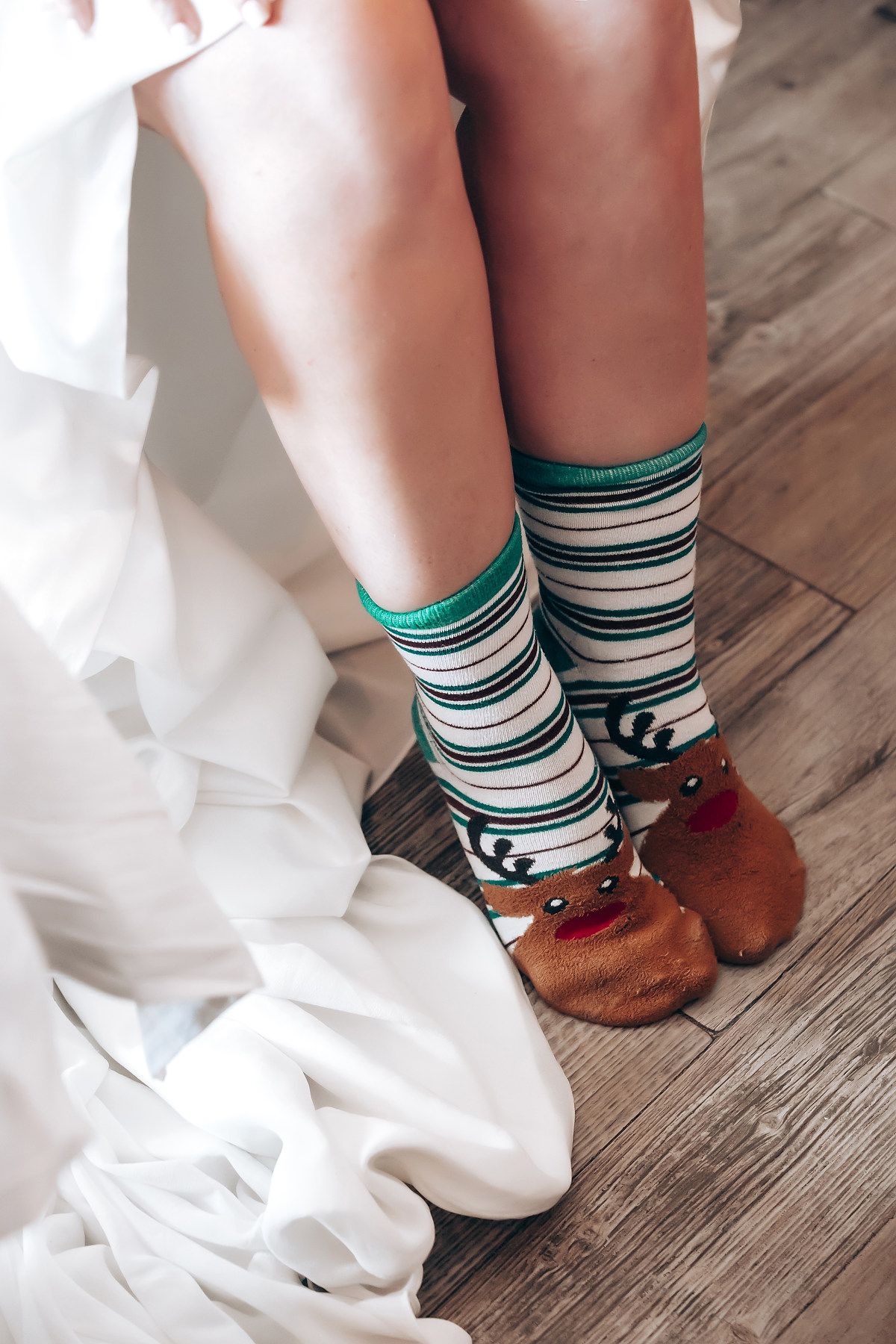 Striped turquoise and white socks with brown reindeer slippers shown on hardwood floor next to white fabric.