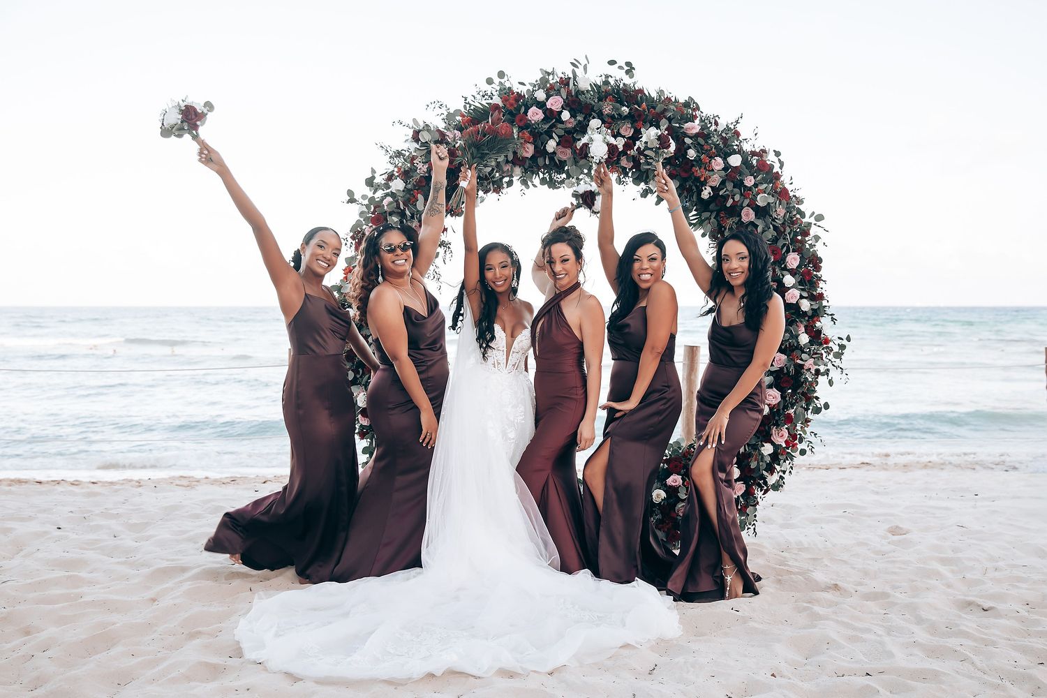 Bridesmaids in burgundy dresses pose with the bride under a floral arch on a tropical beach.