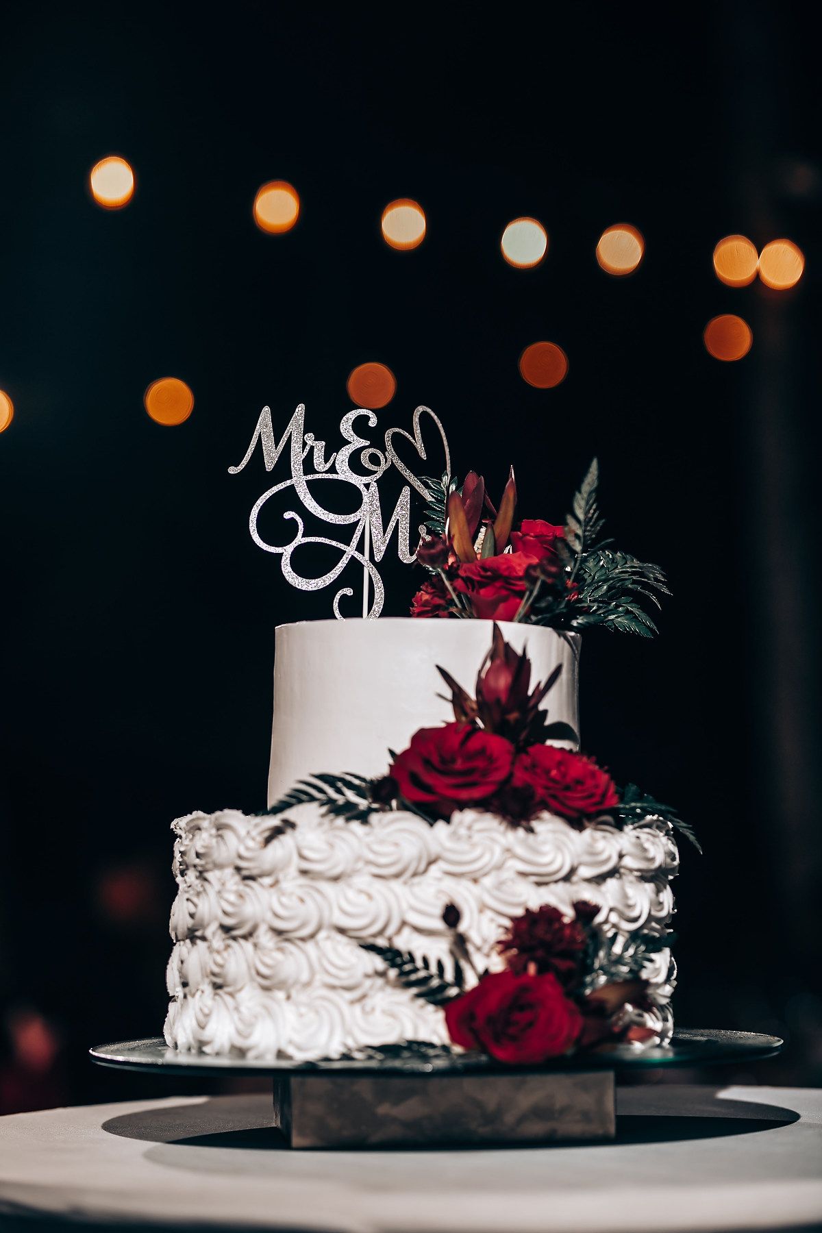 Elegant white wedding cake decorated with deep red roses and custom topper against dark background.