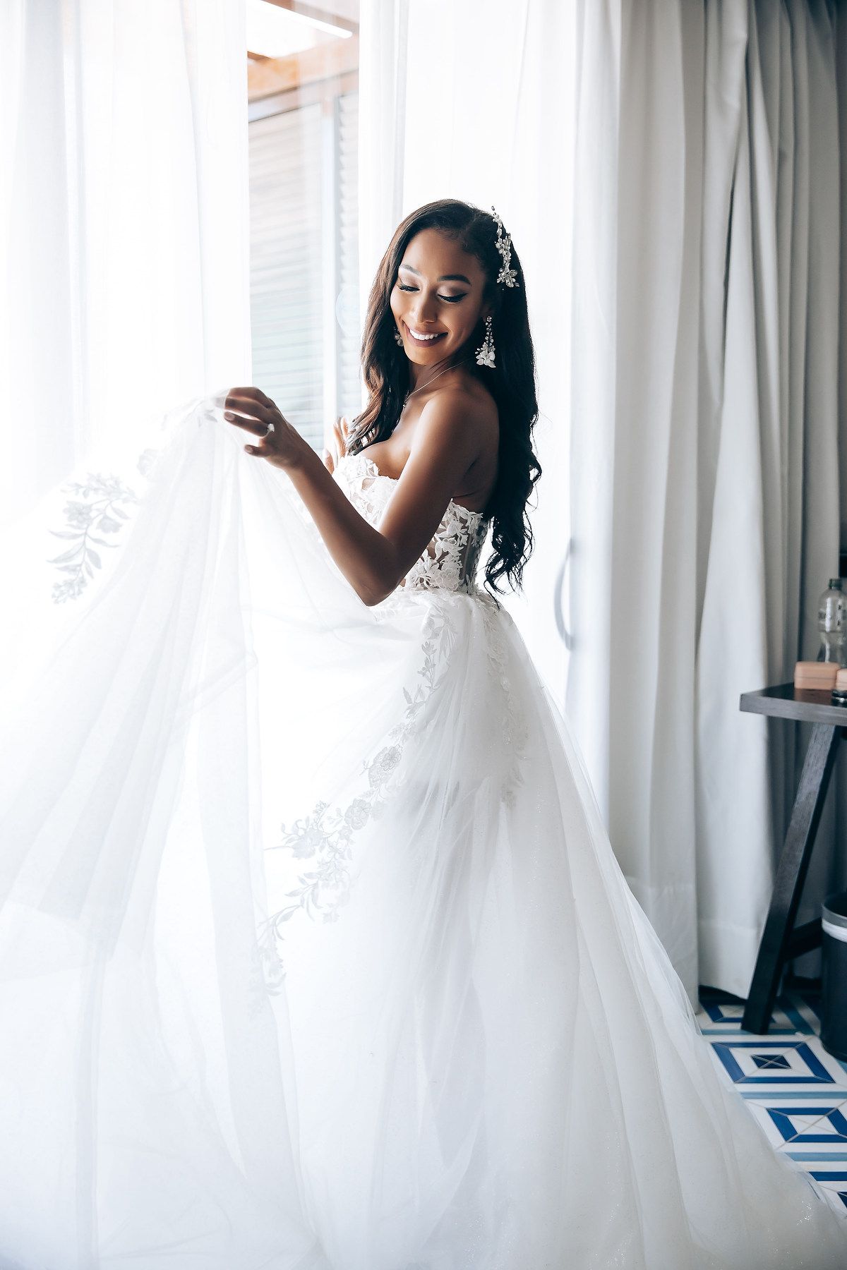 Bride in a flowing white wedding gown twirls playfully by sheer curtains in a bright room.
