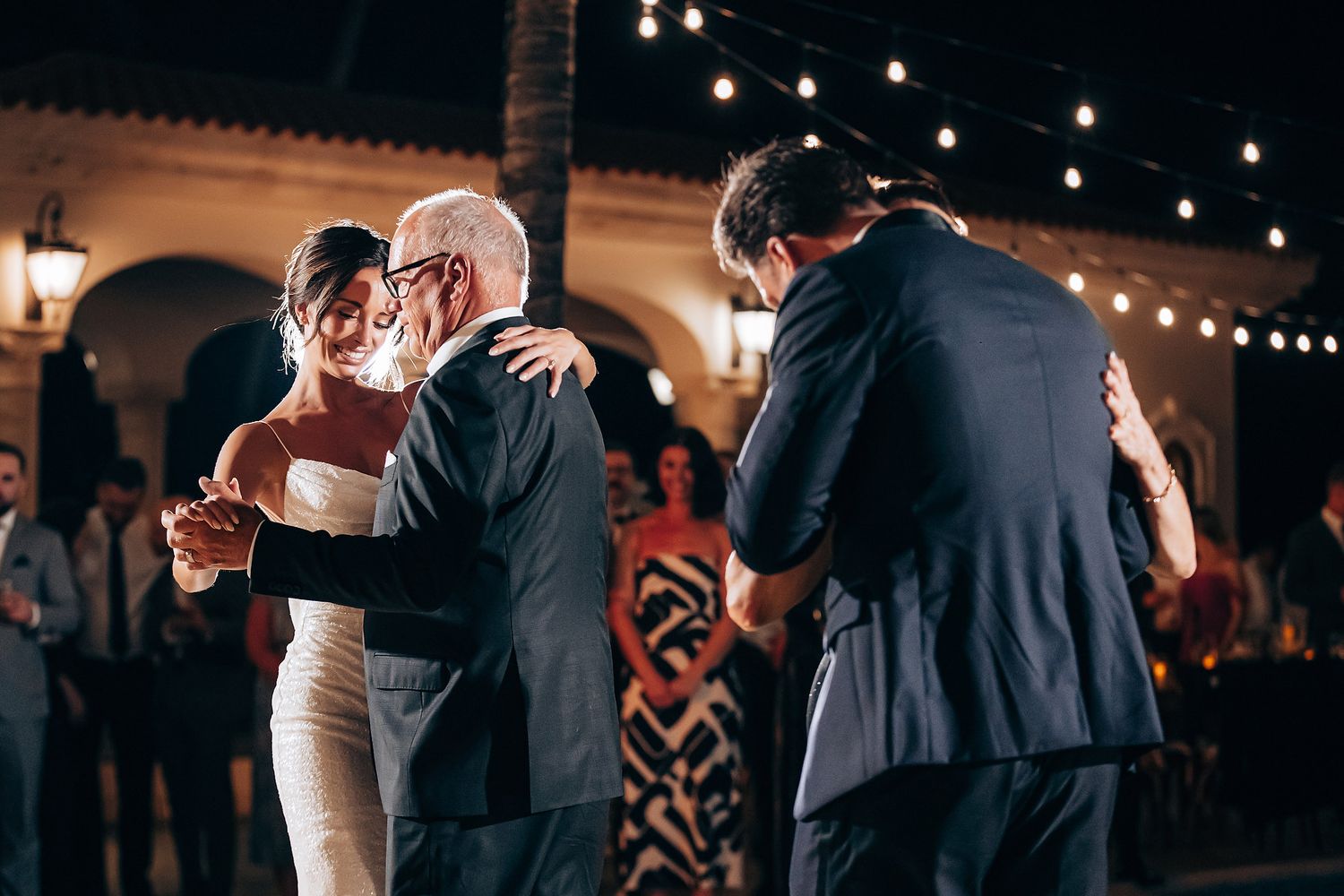 String lights illuminate a romantic wedding dance floor as couples sway together at an evening reception.