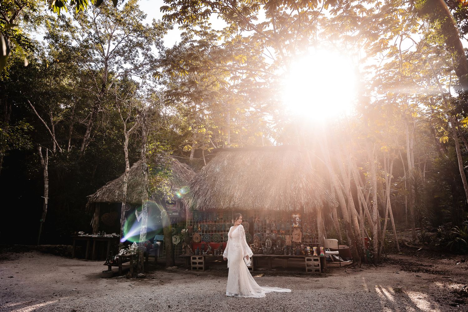 A dreamy wedding scene with sunlight streaming through trees onto a thatched-roof structure.