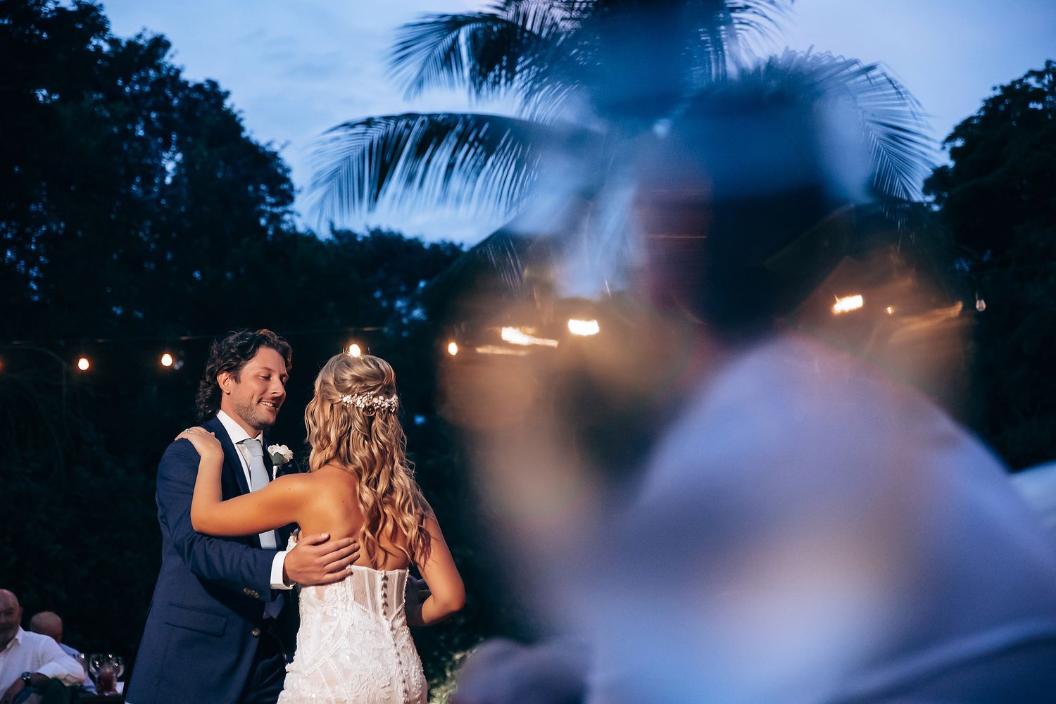 Evening wedding reception under string lights with palm trees silhouetted against blue sky.