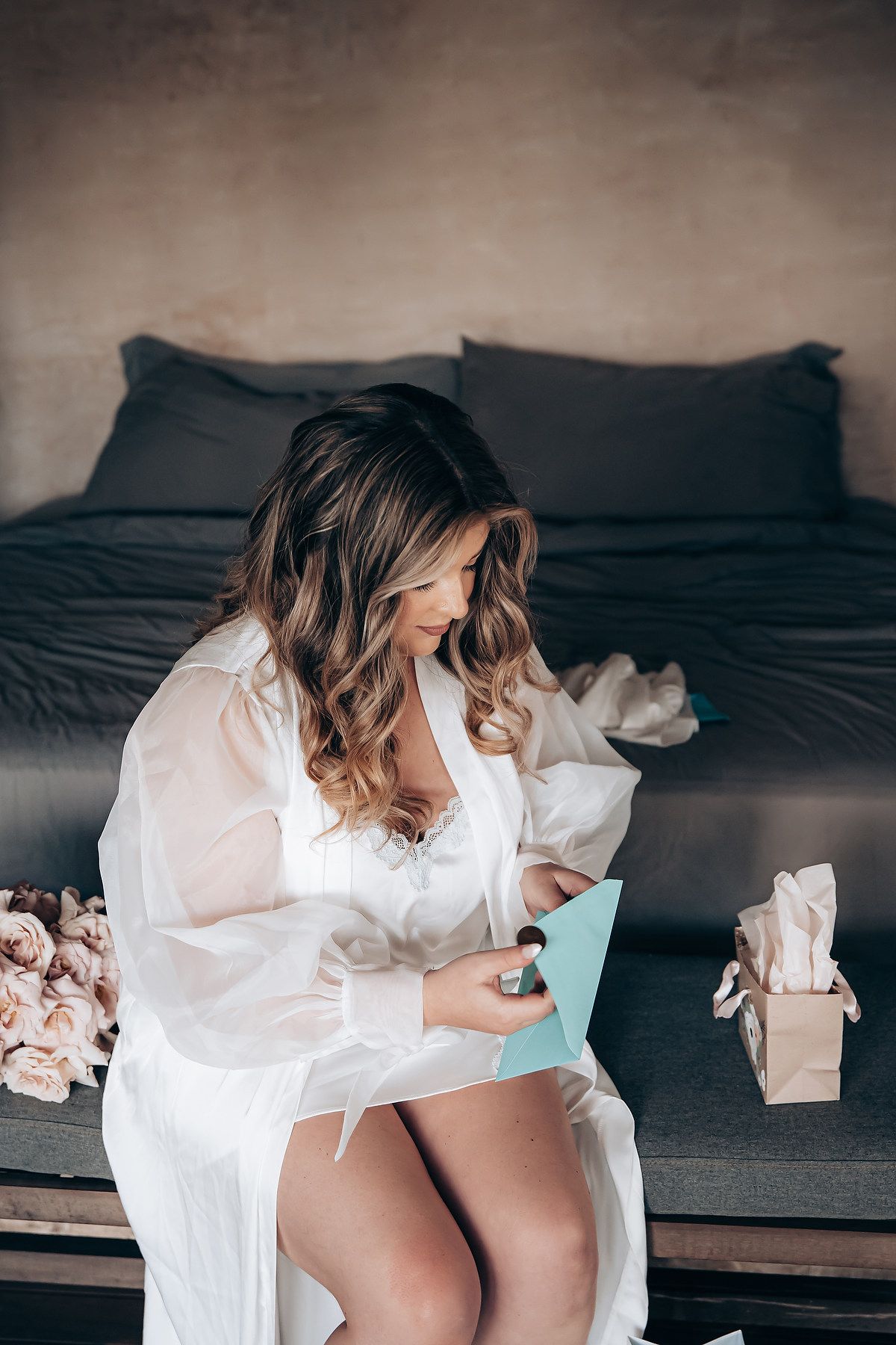 Woman in white flowing dress reclining on dark bedding in moody boudoir photoshoot.