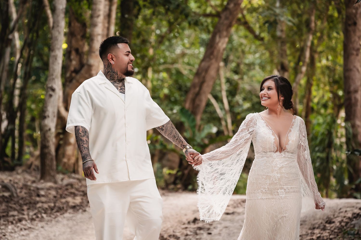 A joyful couple holding hands and laughing together in a forest setting wearing wedding attire.