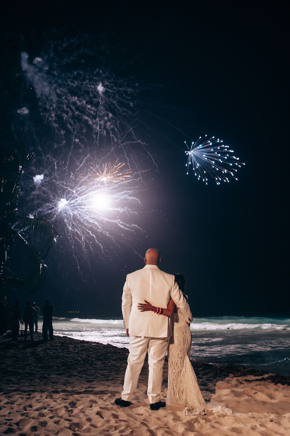 A couple watches fireworks display on a beach at night, silhouetted against the illuminated sky.