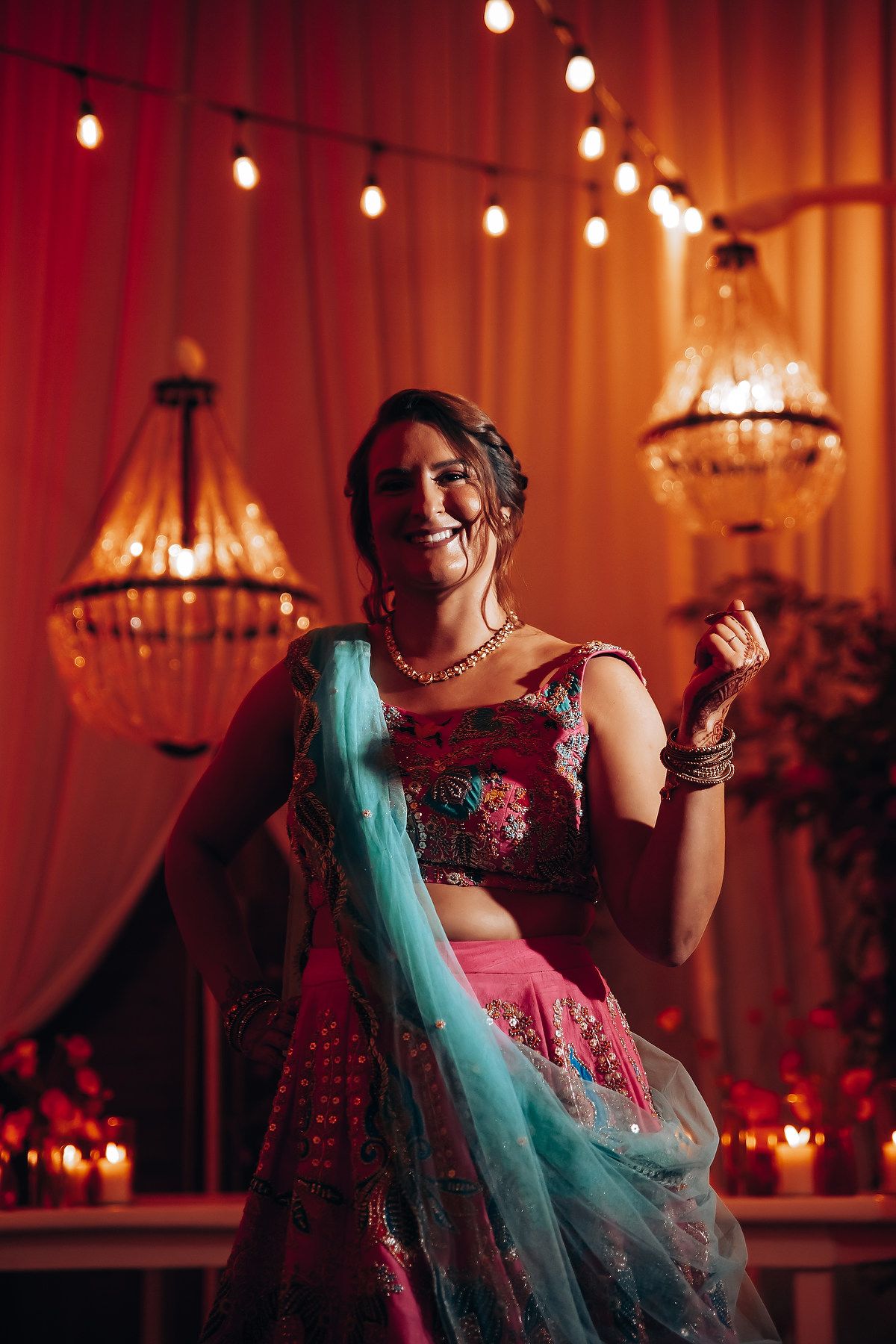 A person in a vibrant turquoise and pink lehenga poses under warm chandelier lighting.