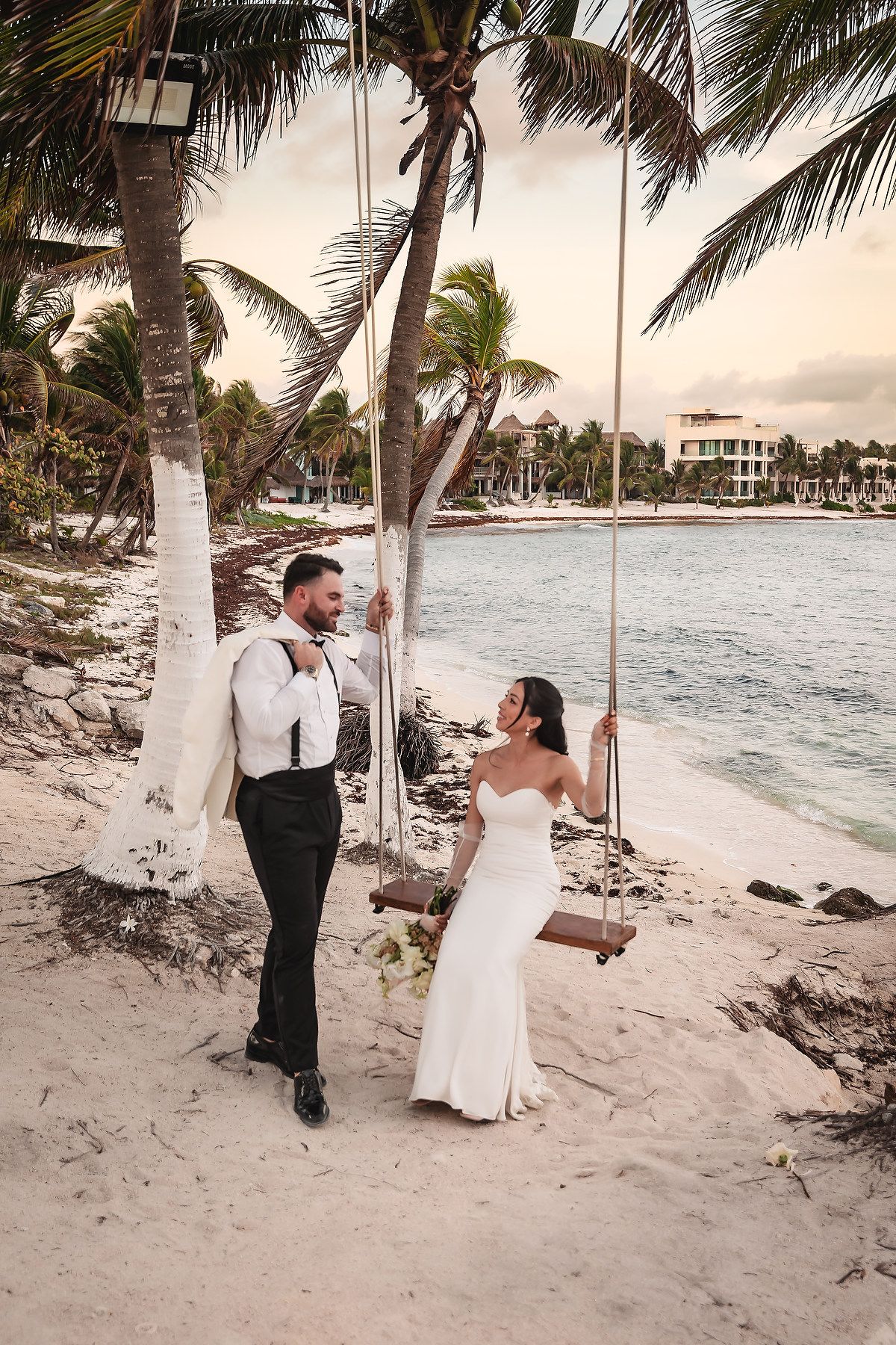 Romantic beach scene with couple by wooden swing under palm trees at sunset.