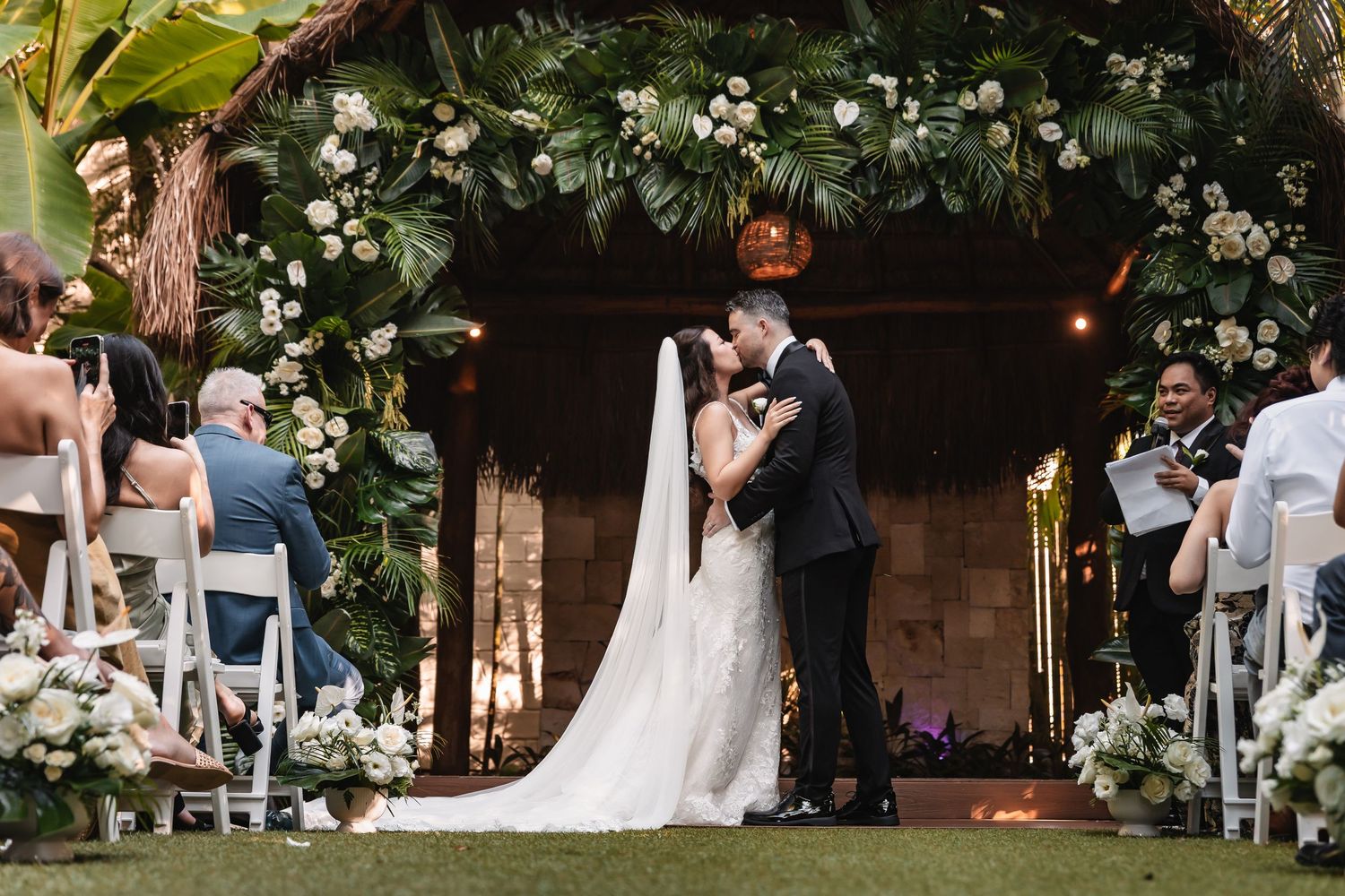Outdoor wedding ceremony setup with white floral arch and guest seating in a garden setting.