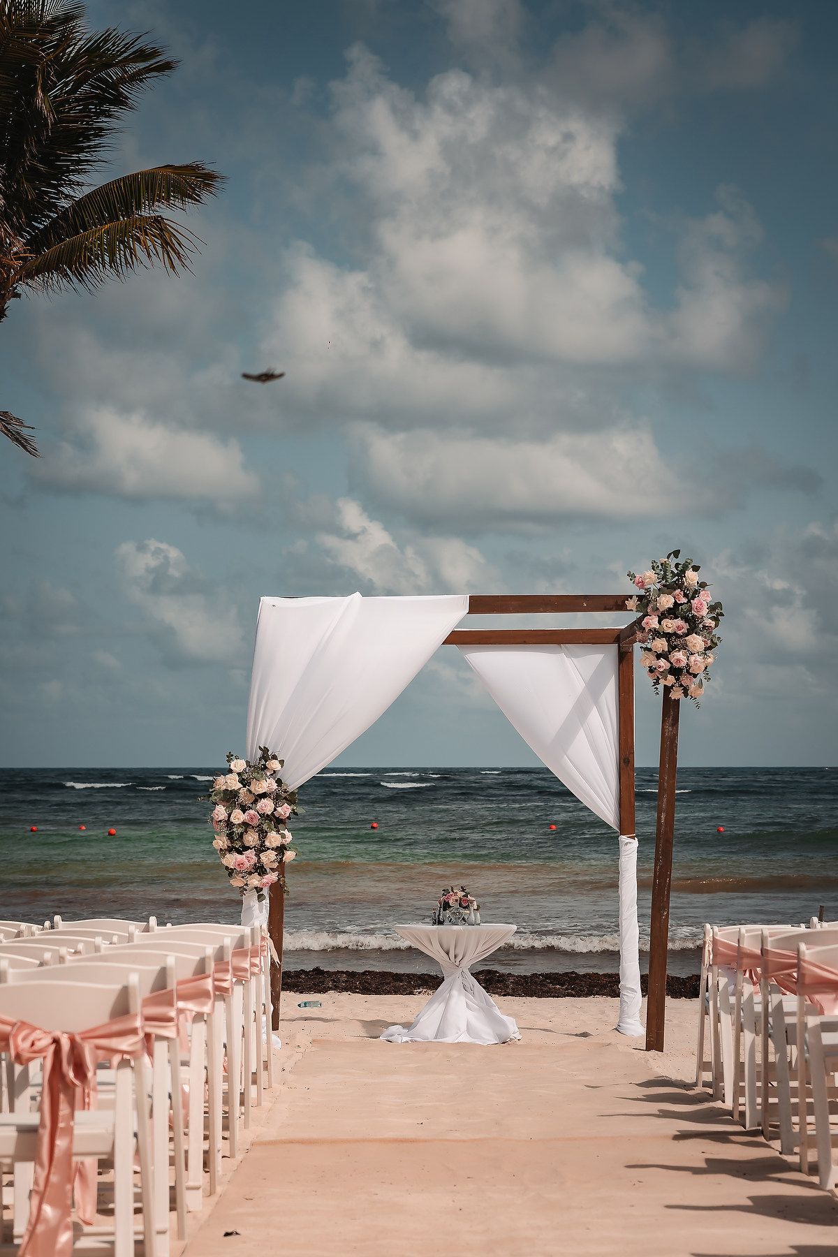 Beachfront wedding ceremony setup with white draped arch and pink floral decorations.