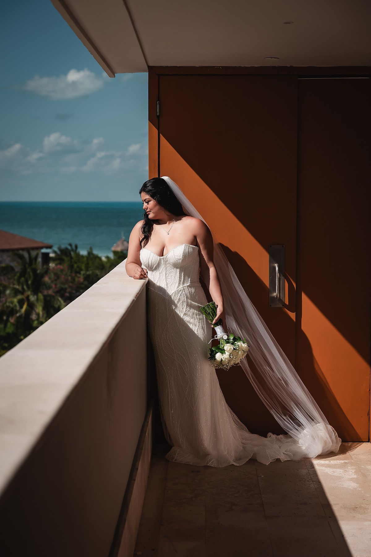 A flowing white wedding dress captured in dramatic lighting on a balcony overlooking the ocean.