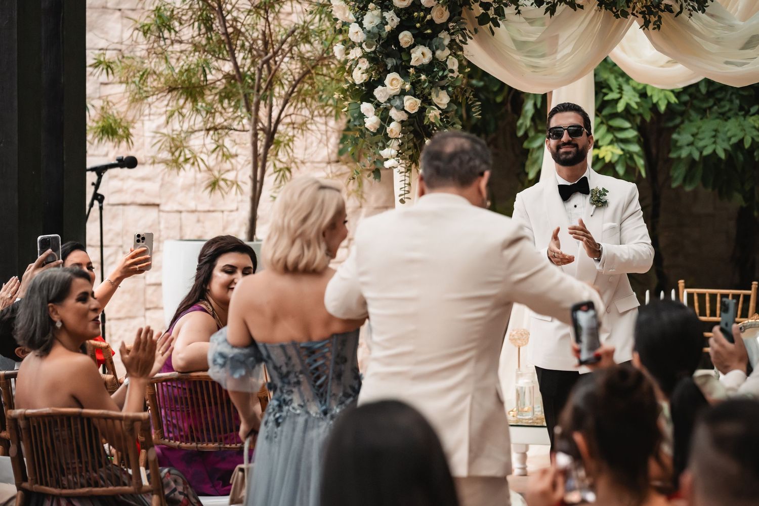 Wedding guests gather under a covered outdoor patio area decorated with white draping and floral arrangements.