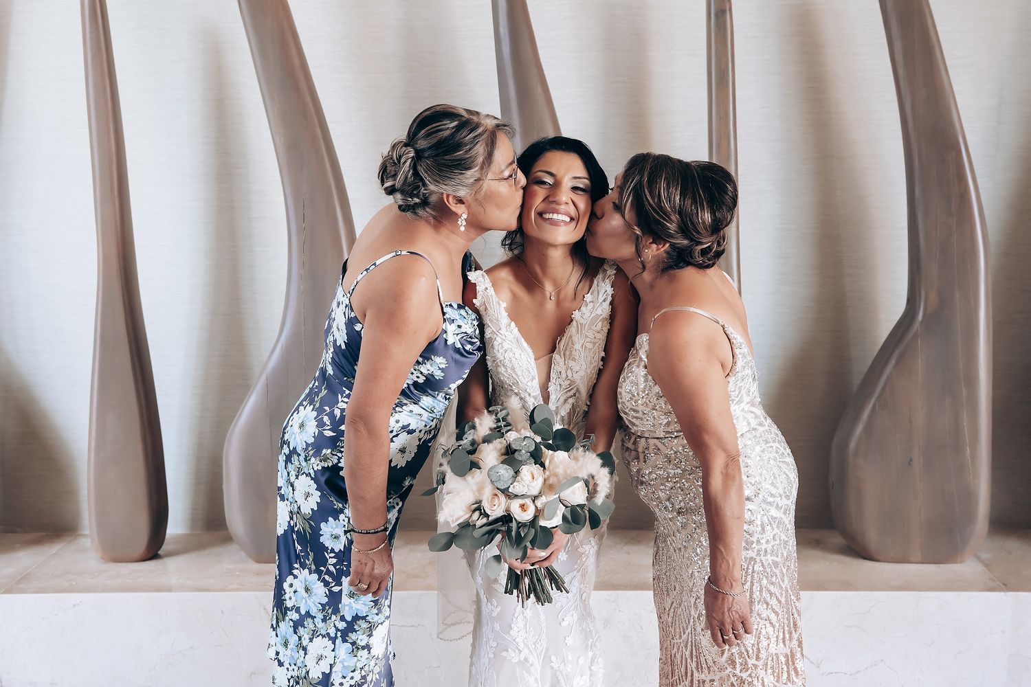 Three women in formal dresses share an intimate moment with floral bouquets against a neutral backdrop.