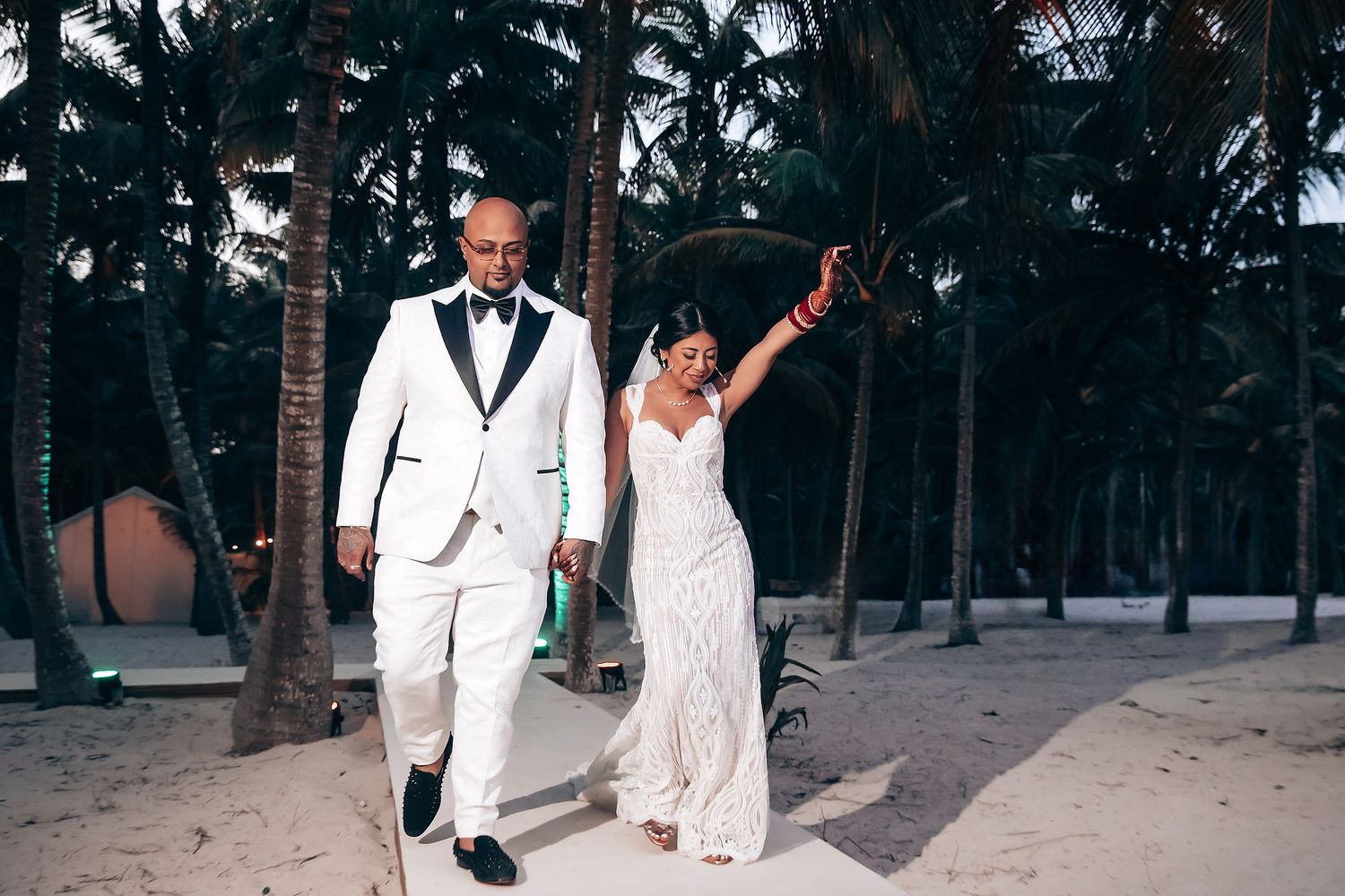 Wedding party in white attire poses for photos on a palm-lined beach at sunset.