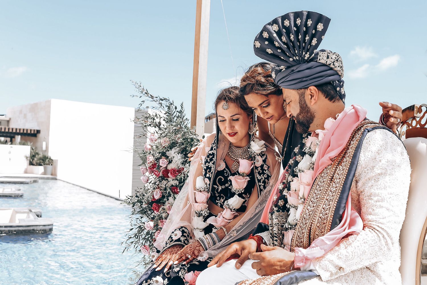 Three people in traditional Balinese headdresses sit together by a pool, sharing a joyful moment.