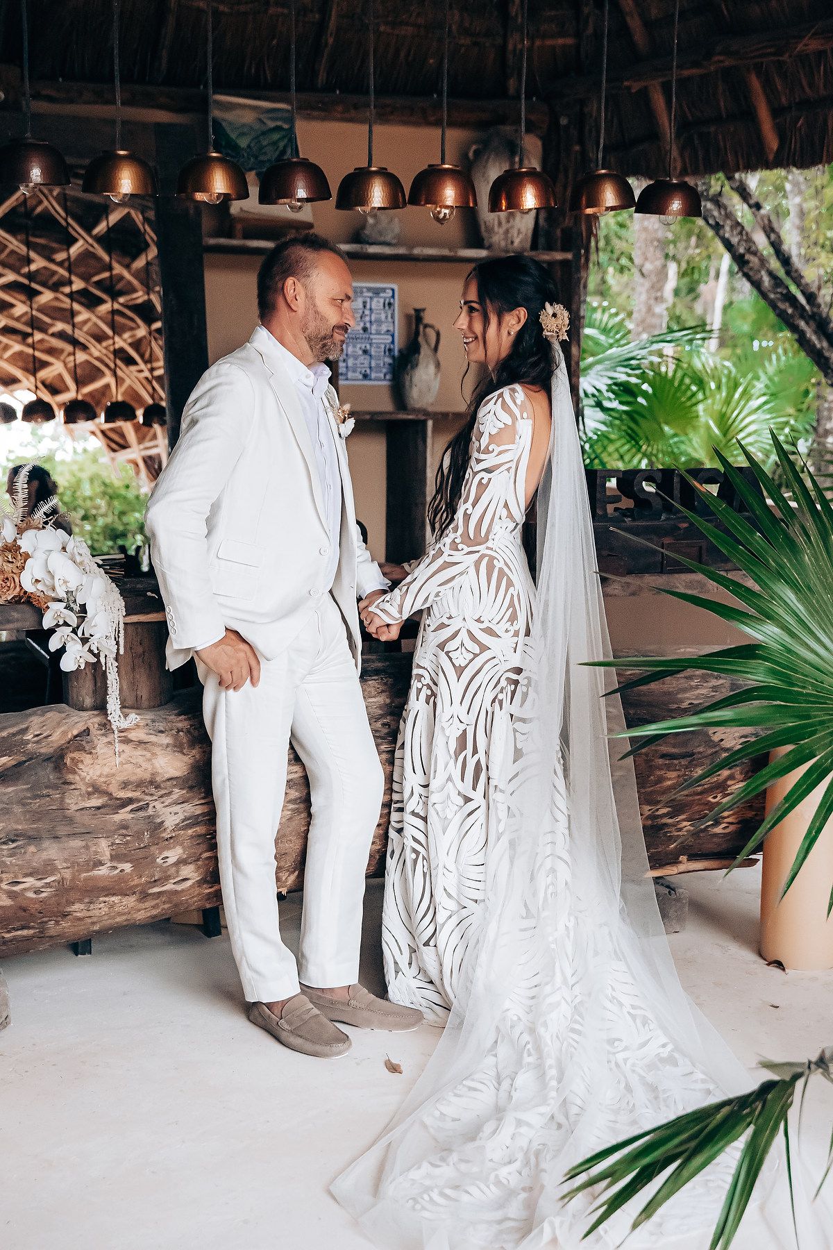 Wedding reception scene with guests in white attire dancing under covered patio with palm decor.