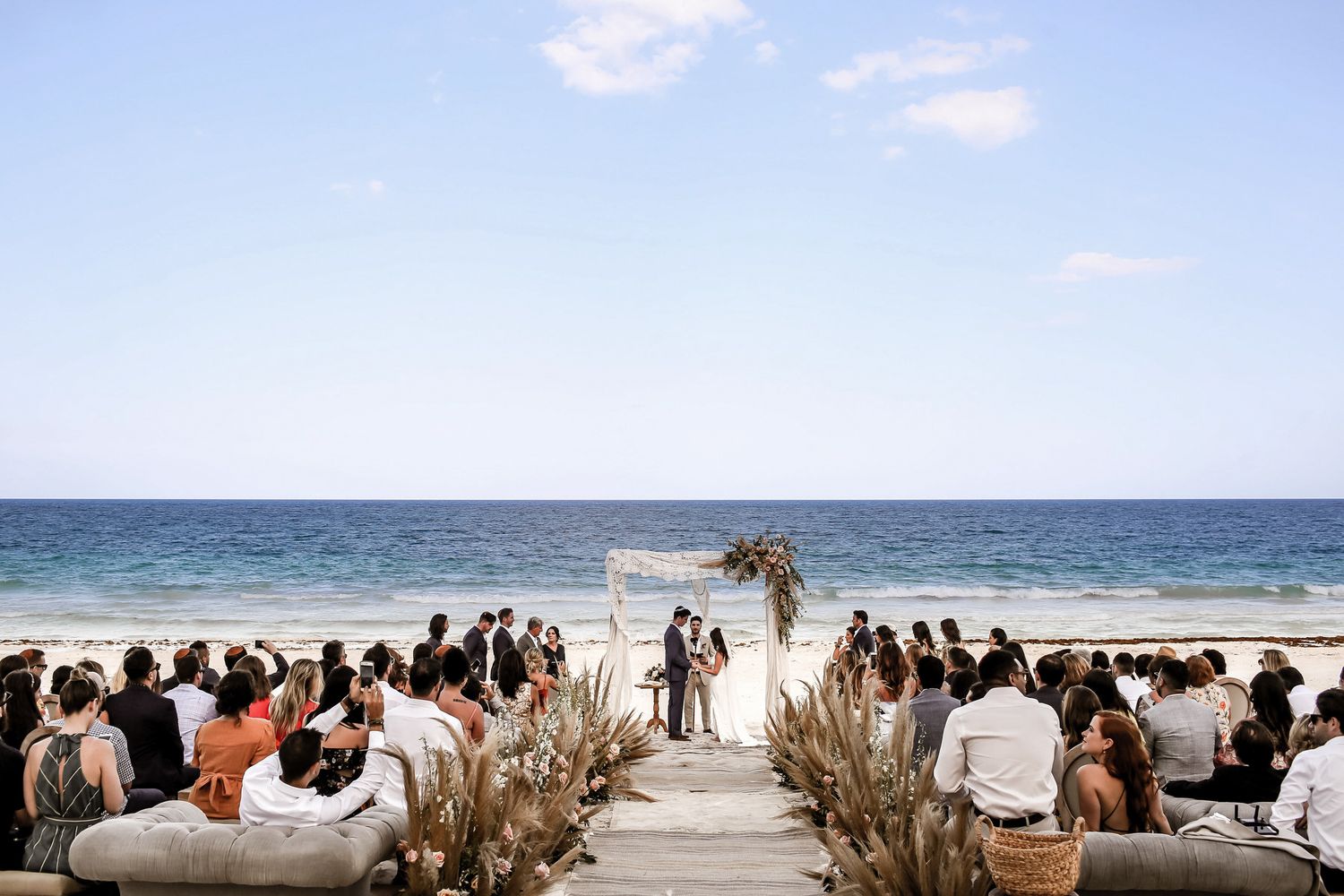Beachfront wedding ceremony setup with dried pampas grass aisle decorations and ocean backdrop.