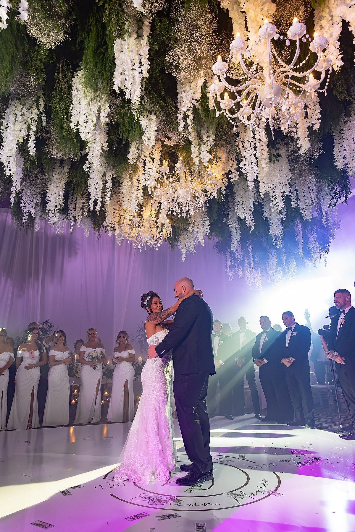 A couple shares their first dance under hanging white floral arrangements and purple lighting.