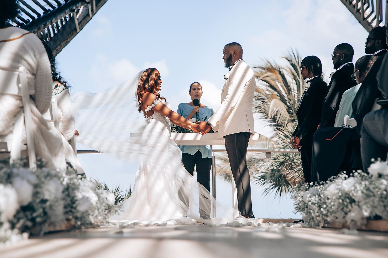Wedding ceremony taking place under a modern geometric structure with tropical beach backdrop.
