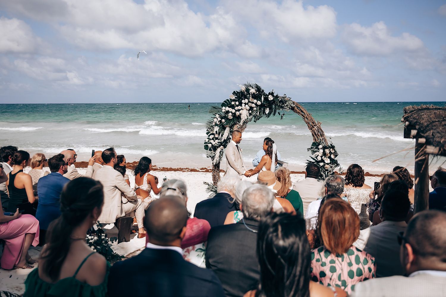 Wedding guests seated on a beach watching a ceremony under a floral arch by the ocean.
