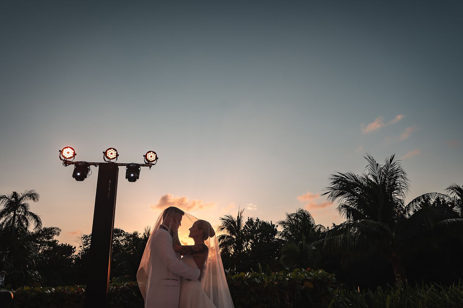 Sunset silhouettes of a wedding ceremony against a dramatic evening sky with palm trees.