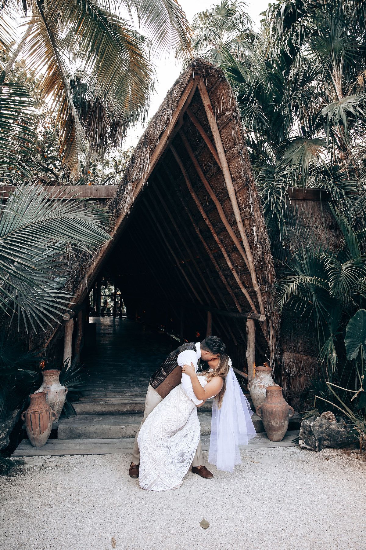 A series of romantic photos showing a couple in white attire posing near a rustic thatched hut surrounded by palm trees.