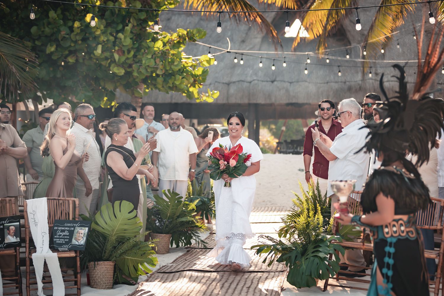 Outdoor beach wedding ceremony with bride holding red bouquet, guests applauding, string lights, and tropical greenery.