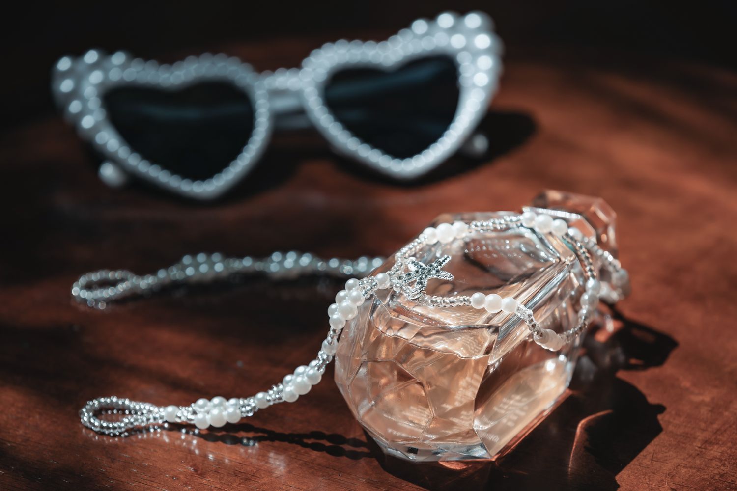 Heart-shaped rhinestone sunglasses beside a pearl-adorned perfume bottle on a dark wooden surface.