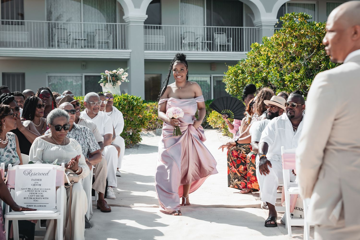 Bridesmaid in pink gown walks down outdoor wedding aisle at beachside resort ceremony with seated guests.