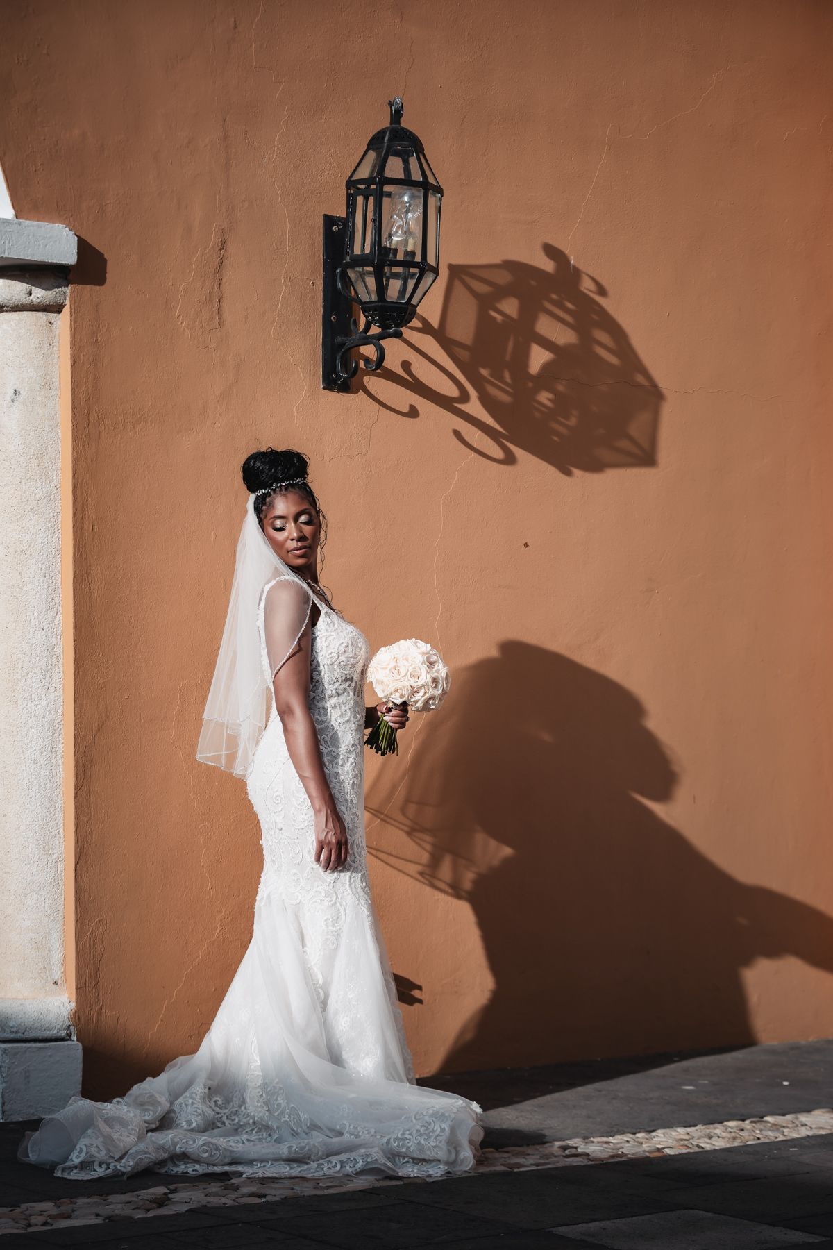 Elegant bride in white lace mermaid gown holding bouquet, posing against warm terracotta wall with ornate lantern.