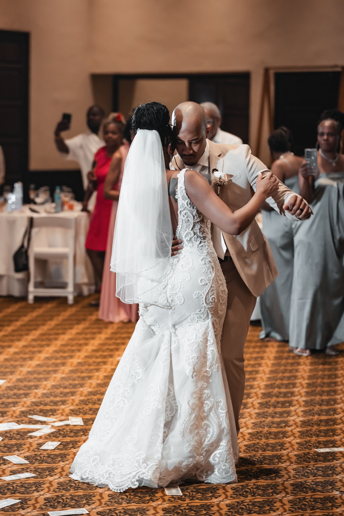 Bride and groom share their first dance at wedding reception, bride in lace gown with veil, groom in tan suit.