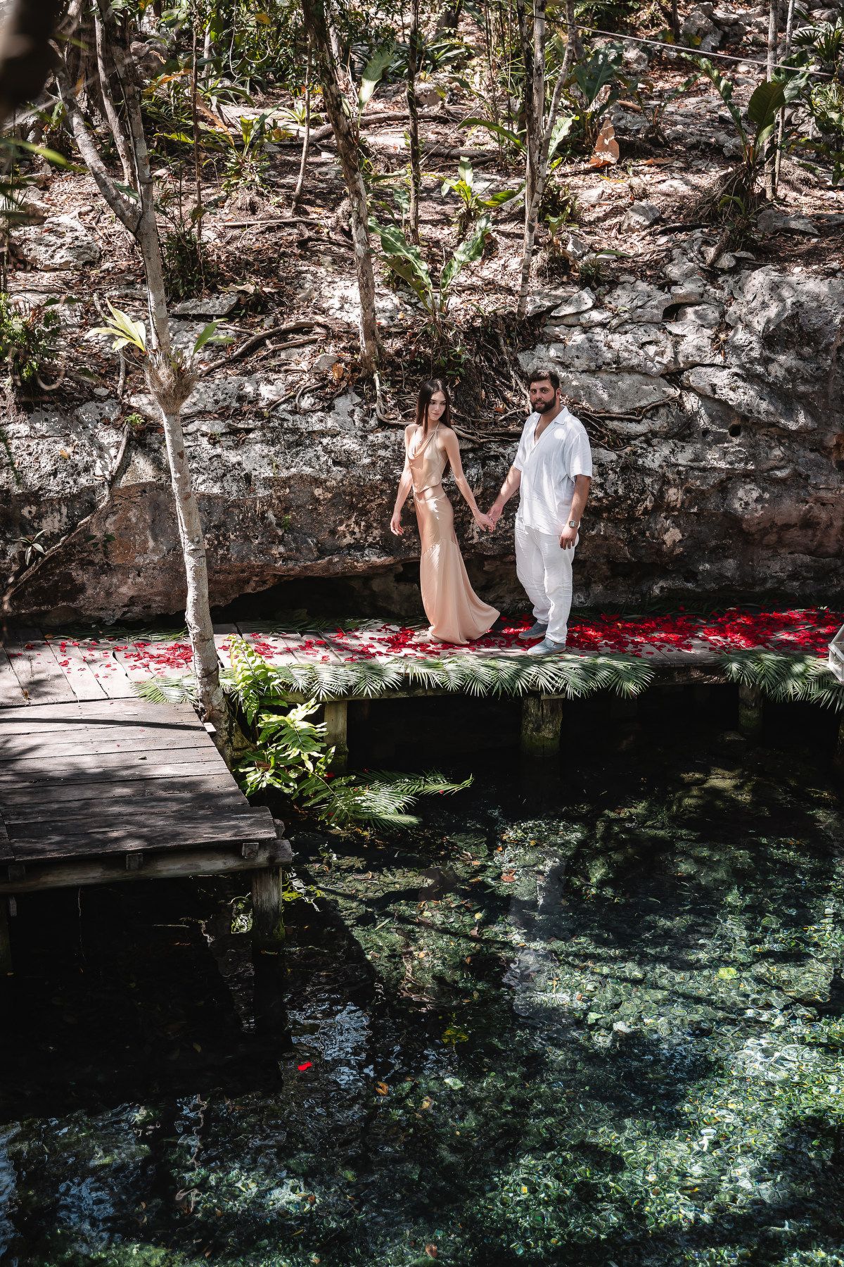 Couple holding hands on a flower-petal-covered dock over a crystal-clear cenote surrounded by lush jungle.