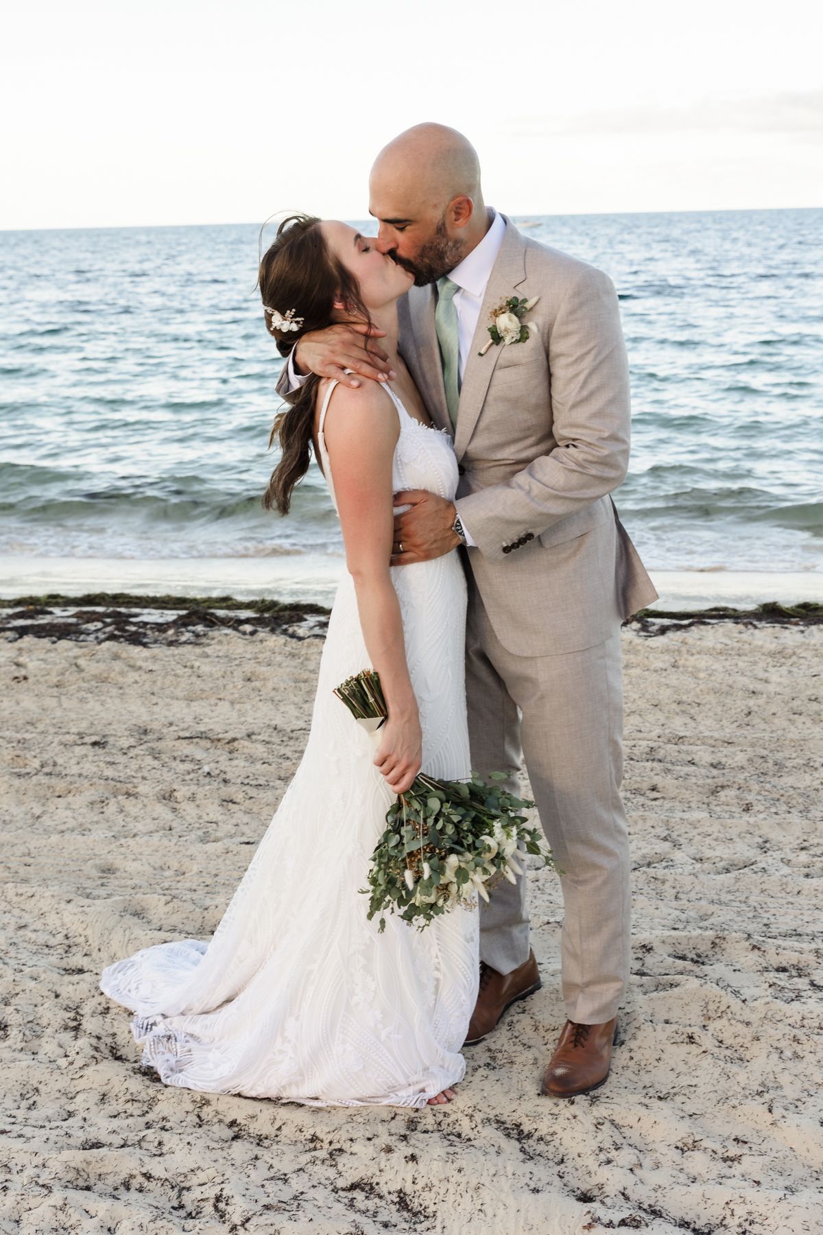 Bride and groom share a romantic beach kiss, she holds a bouquet in a lace gown as he wears a tan suit.